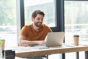 Smiling businessman using laptop near coffee and eyeglasses on table in office