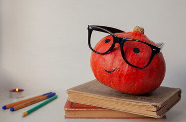 Pumpkin as a student in hipster glasses on books with pens and pencils. Clever pumpkin at school for Halloween in autumn. Nerd pumpkin with glasses.