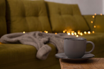 Gray cup of tea on the wooden table against green home sofa and warm garland lights.Empty space