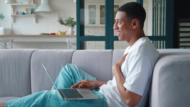 Side portrait of a friendly African American guy resting at home, and having a video call on laptop. Handsome young man talking to his interlocutor on laptop. Online communications concept