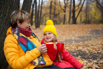 grandmother and little granddaughter sit and  drinking hot tea from thermos in autumn park. Happy family enjoying picnic outdoors.