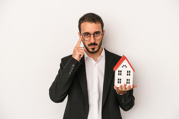 Young real estate agent man holding a model house isolated on white background pointing temple with finger, thinking, focused on a task.
