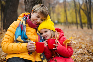 grandmother and little granddaughter sit and  drinking hot tea from thermos in autumn park. Happy family enjoying picnic outdoors.