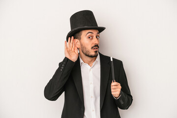 Young magician man holding wand isolated on white background trying to listening a gossip.