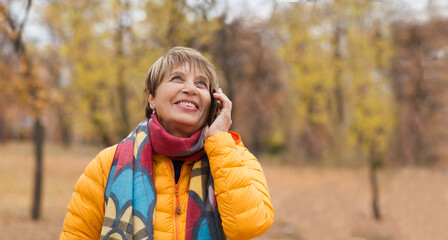 happy senior woman in a scarf and jacket uses a smartphone in the autumn park.  