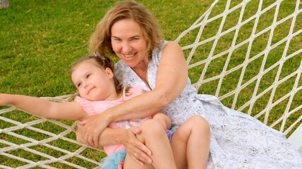 funny couple grandmother and granddaughter 3 years old are relaxing and basking in a hammock on a green meadow in their garden on a summer sunny day. Vacation concept, generational relationship