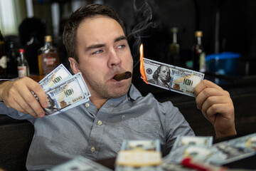 Man lighting his cigar with 100 dollars banknote against background of bottles in the bar. The...