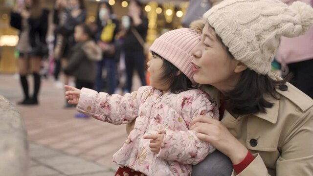 taiwanese mother and baby smiling with joy while having fun watching dancing fountain on a city square with group of tourists taking photos at background