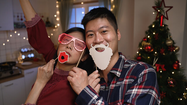 funny asian couple looking at mobile cam is playing with xmas masks on stick while they are having fun taking selfies with Christmas tree on background at home