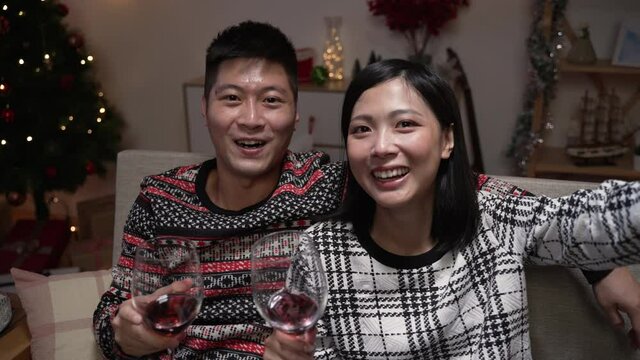 joyful Taiwanese young couple holding wineglasses and talking happily to mobile camera while having a video call with friends in the living room on Christmas eve