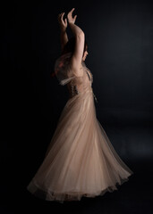Full length  portrait of red haired  girl wearing a creamy fantasy gown like a fairy goddess costume.  standing  pose with elegant gestural movement  backwards the the camera, isolated on dark studio 