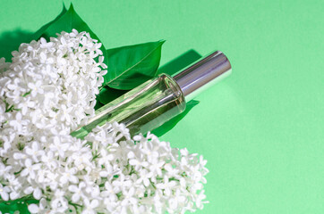 White lilac flowers and a glass bottle with perfume on a green background. Bright shadows and light. Place for your text. Flat lay.
