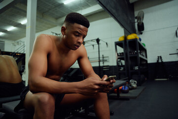 Fototapeta na wymiar Young African American male looking at his phone to reply to an online message. male personal trainer looking at his phone while sitting down in the gym. High quality photo 