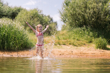 Caucasian little girl in bright swimming suit playing splashing jumping in river in hot summer day. Full length horizontal shot. Green blurred background. Ecolife and happy summertime concept.