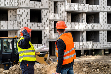young strong hardy construction workers in overalls standing near a new building with a plan