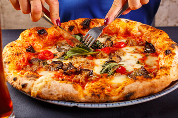 Pizza napoletana con sugo di pomodoro, salsiccia, basilico fresco e melanzane fritte mentre viene tagliata con forchetta e coltello