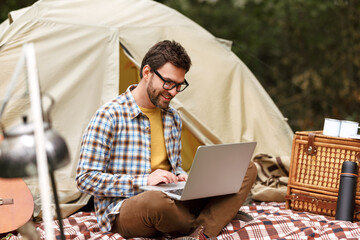 Smiling man sitting near tent with laptop computer and working remotely while camping in nature