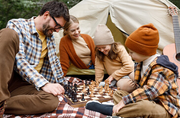Happy smiling family playing chess game at campsite during camping trip in nature