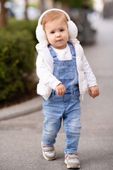 Cute baby girl 1-2 year old wear fluffy white headphones, denim suit pants and knitted sweater posing on city street outdoors. Childhood. Happy little child.