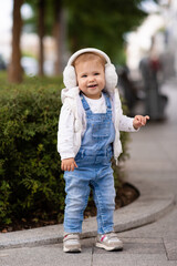Fototapeta premium Cute baby girl 1-2 year old wear fluffy white headphones, denim suit pants and knitted sweater posing on city street outdoors. Childhood. Happy little child.