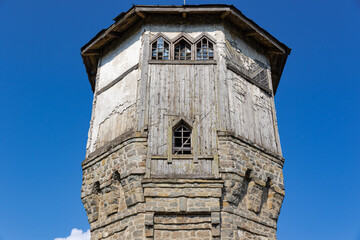 Obraz premium The upper part of an ancient watchtower made of stone blocks and wood paneling with windows against the blue sky on a bright sunny summer day. Close-up