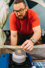 A young bearded happy man sitting in his tent on a camping trip and preparing coffee on a little electric stove.