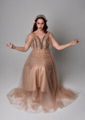 Full length  portrait of red haired  girl wearing a creamy fantasy gown and crystal crown, like a fairy goddess costume.  sitting pose with elegant gestural hands, isolated on light studio background.