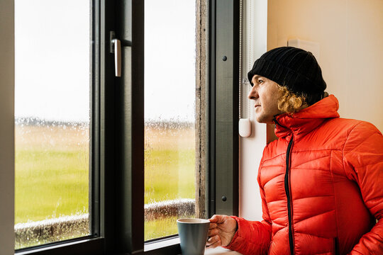 Dreamy guy standing near window in kitchen and drinking coffee