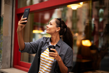 Happy girl enjoying in sunny weather and fresh coffee. Gorgeous casual brunette drinking coffee to go. Young woman having video call