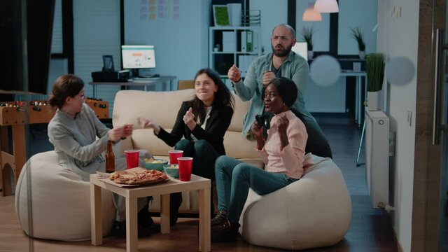 Happy colleagues playing video games on console television with controllers after work at office. Workmates dancing after winning game with joysticks on tv, enjoying fun activity.