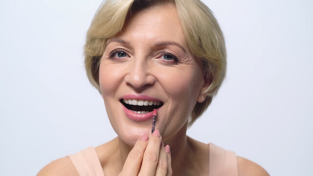 Happy Middle Aged Woman Applying Lip Gloss While Looking At Camera Isolated On White.