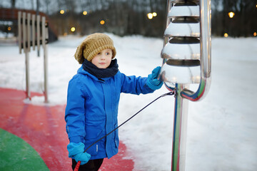 Little boy knocking by street xylophone drums on playground in public park on a snow winter day....