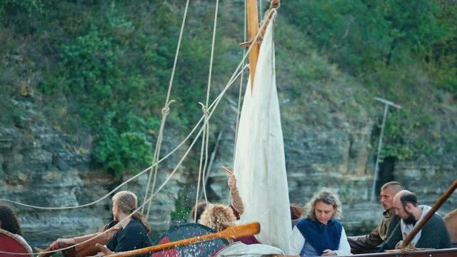 Vikings Sail on an Old Ship with a Lowered Sail on a Quiet River Against the Backdrop of Rocky Mountains. Concept on the theme of the Vikings and the early Middle Ages.