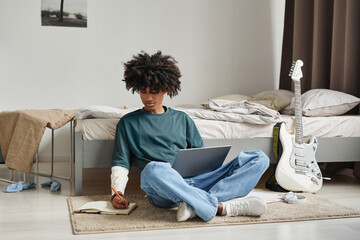 Full length portrait of teenage African-American student sitting on floor at home or in college...