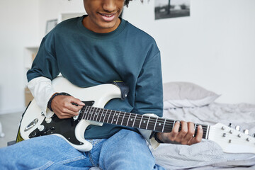 Cropped portrait of smiling African-American teenager playing guitar while sitting on bed at home,...