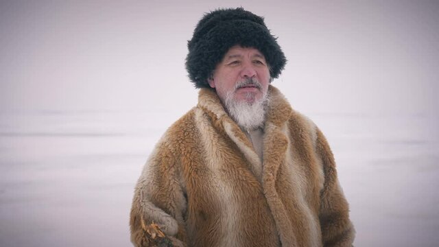 Zoom in indigenous old man standing at picturesque winter landscape looking away thinking. Handsome confident senior adventurer looking at camera admiring beauty of nature outdoors. Slow motion