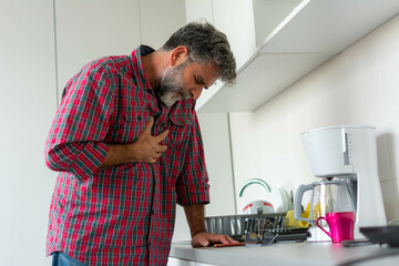 Senior man with grey beard suffering from chest pain in kitchen at home. Man with chest pain suffering from heart attack at home. Elder Man With Chest Pain.