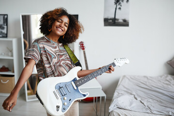 Waist up portrait of smiling African-American teenage girl playing electric guitar at home, copy...