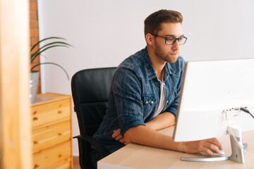 Side view of focused young man wearing stylish glasses working on desktop computer sitting at desk at home office in room with modern interior. Freelance programmer working from apartment.