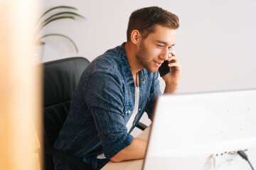 Side view of focused young business man talking on mobile phone sitting at desk with monitor computer at home office. Close-up of handsome businessman having conversation on smartphone at workplace.