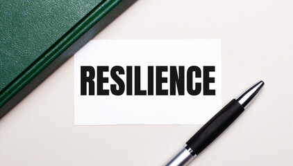 On a light gray background lies a pen, a green notebook and a white card with the text RESILIENCE. Business concept.