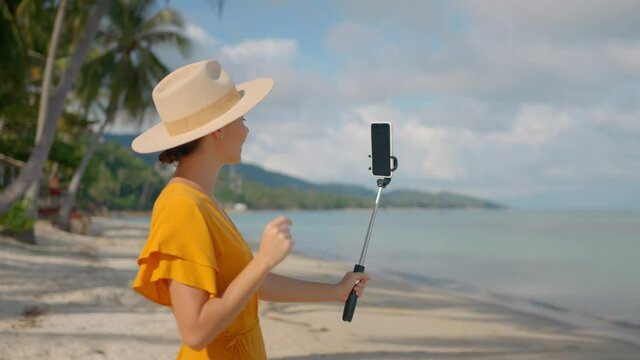 Beautiful girl in the straw hat doing online report on the ocean beach holding phone on monopod 
