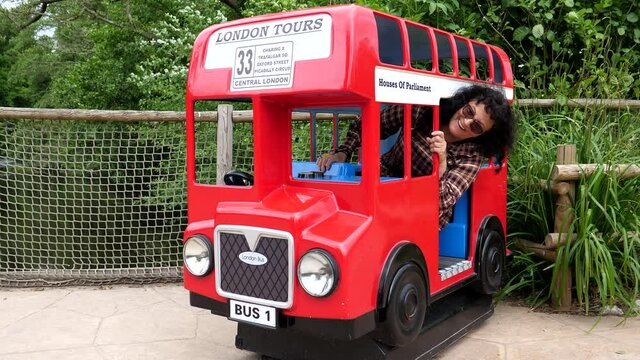 Adult woman sitting inside of small red British bus for children's entertainment, like Alice in wonderland, is having  a fun, laughing and waving her hand.