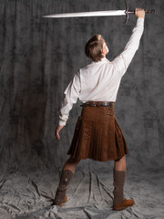A young man in a leather kilt and a white lace-up blouse. He raised the weapon over his head. A Scottish knight with a two-handed sword. Full-length Photo in the studio back view, on a gray background