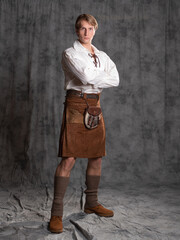 A young man in a leather kilt and a white lace-up blouse. A Scottish, Full-length Photo in the studio on a gray background