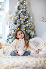 Happy little smiling girl with gift on Christmas Eve sit on bed. Child opens New year gift near christmas tree with glowing lights garland. Kid play in bed with xmas balls and toys in bedroom at home	