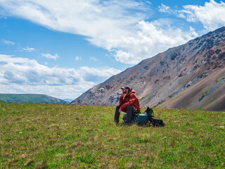 Fototapeta na wymiar Tired tourist sits on a large backpack and happily looks into the distance against the background of blue dramatic clouds against the background of tundra mountains. Rest time on a hike, a rest stop.