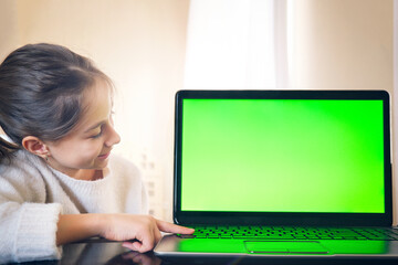 A young girl looks at the green screen of a laptop and presses her finger on the keys.