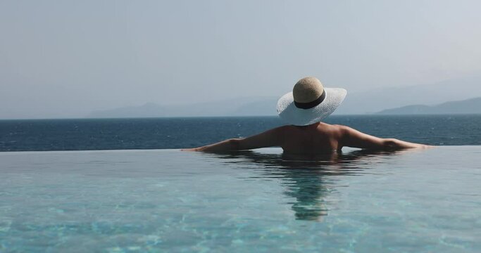 woman with straw hat relaxing in infinity swimming pool and enjoying sea view at luxury resort
