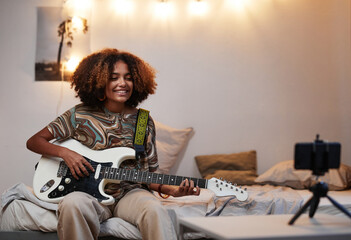 Portrait of young African-American woman playing guitar at home and recording video or...